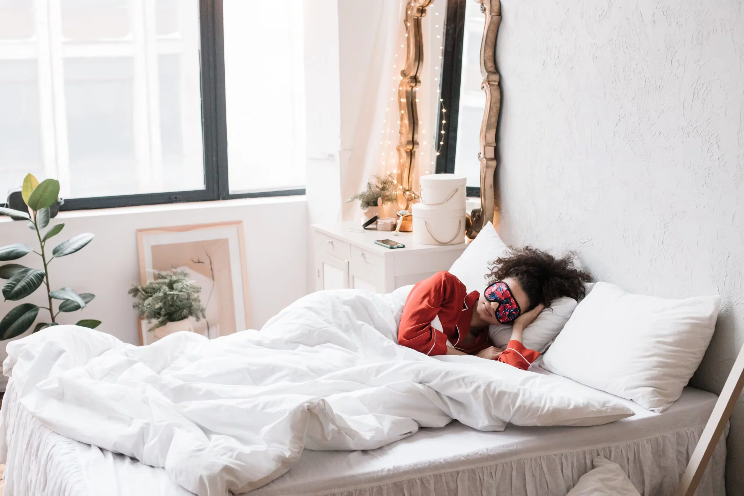 Woman wearing a sleep mask, resting on a cozy white bed surrounded by indoor plants in a bright bedroom.